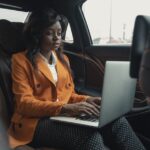 Focused businesswoman wearing an orange blazer and working on a laptop inside a car, showcasing productivity.