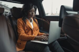 Focused businesswoman wearing an orange blazer and working on a laptop inside a car, showcasing productivity.