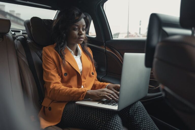Focused businesswoman wearing an orange blazer and working on a laptop inside a car, showcasing productivity.