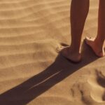 Close-up of a person standing barefoot on sunlit sand with visible footprints.