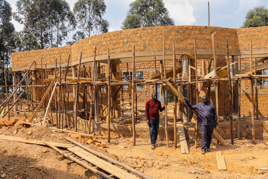 Two men inspect the wooden scaffolding of a brick building construction site outdoors.