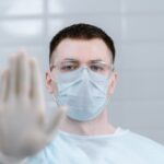 Healthcare worker wearing protective gear and mask making stop gesture in hospital setting.