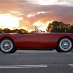 Vintage red convertible car parked on a country road at sunset with dramatic skies.