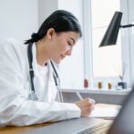 A female doctor writes notes at her desk with a stethoscope around her neck.