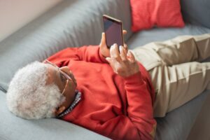 Elderly man in red sweater relaxing on a gray sofa, using a smartphone.