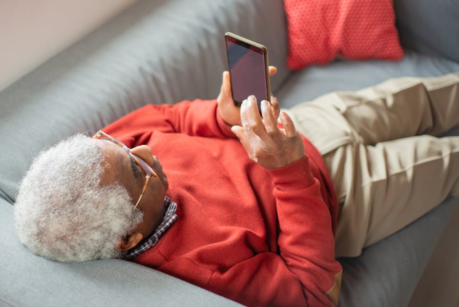 Elderly man in red sweater relaxing on a gray sofa, using a smartphone.