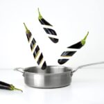 Creative image of sliced eggplants floating above a stainless steel pan against a white background.