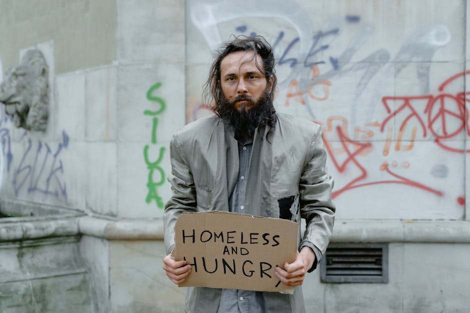 Homeless man standing on the street holding a sign, with graffiti in the background.