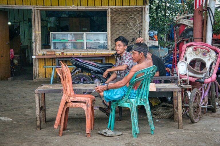 Ethnic males smoking cigarettes sitting near wooden bench and hut in poor Asian town