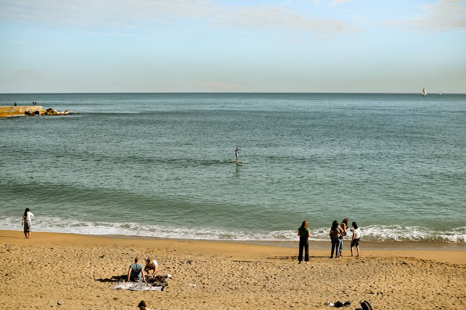 A diverse group of people relax and enjoy leisure time on a sunny beach, with a paddleboarder in the distance.