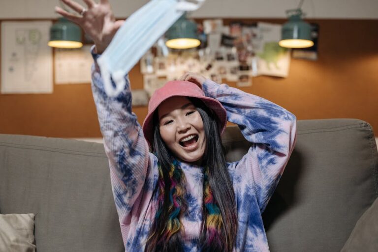 Smiling Asian woman in colorful hoodie and hat joyfully tossing a face mask indoors. Vibrant and lively atmosphere.
