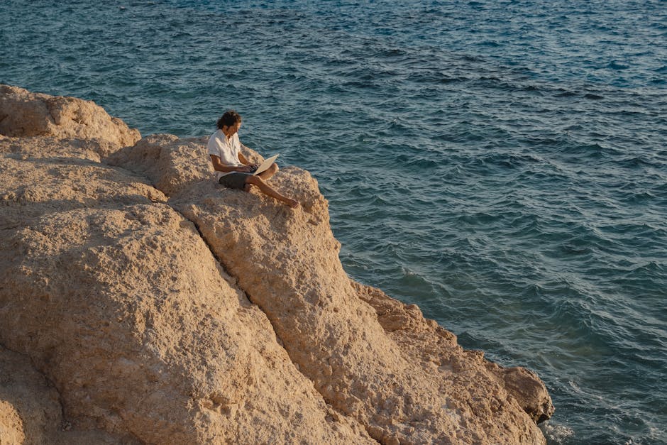 A man sits on a rocky cliff by the sea, working remotely on a laptop.
